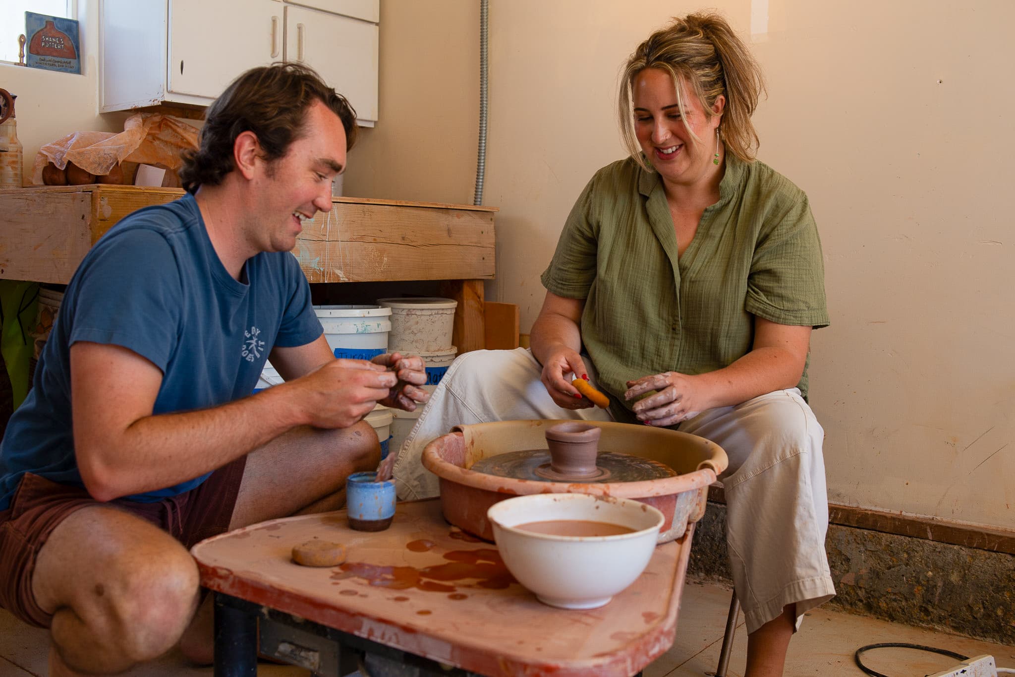 Shane teaching a student on the pottery wheel, both laughing